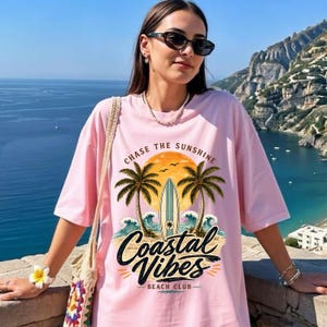 May include: Pink t-shirt with a beach-themed graphic. The design includes palm trees, a surfboard, and the words "Coastal Vibes Beach Club" and "Chase the Sunshine". The shirt has short sleeves and a relaxed fit.