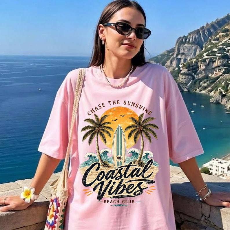 May include: Pink t-shirt with a beach-themed graphic. The design includes palm trees, a surfboard, and the words "Coastal Vibes Beach Club" and "Chase the Sunshine". The shirt has short sleeves and a relaxed fit.