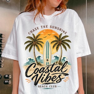 May include: White t-shirt with a beach-themed graphic. The design features a sunset, palm trees, a surfboard, and ocean waves. Text includes "CHASE THE SUNSHINE" and "Coastal Vibes Beach Club".