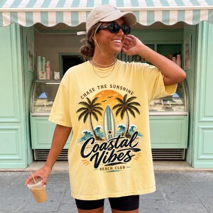 May include: Yellow t-shirt with a graphic that says "Chase the Sunshine" and "Coastal Vibes Beach Club". The graphic includes palm trees, a surfboard, and ocean waves. The shirt is worn with black shorts and a beige cap.