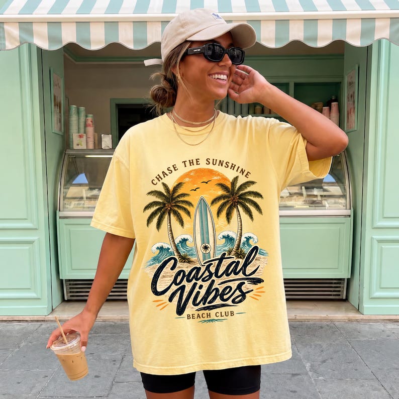 May include: Yellow t-shirt with a graphic that says "Chase the Sunshine" and "Coastal Vibes Beach Club". The graphic includes palm trees, a surfboard, and ocean waves. The shirt is worn with black shorts and a beige cap.