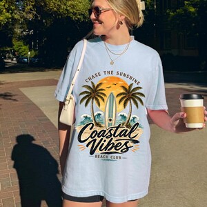 May include: Light blue t-shirt with a coastal graphic. The design features palm trees, a surfboard, and the text "Chase the Sunshine" and "Coastal Vibes Beach Club". The shirt is a relaxed fit.