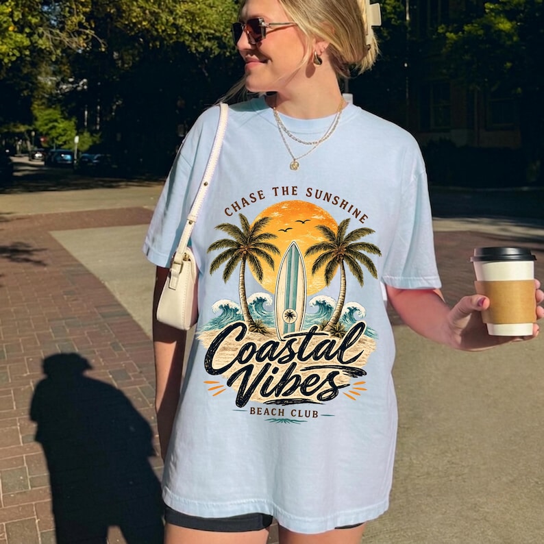 May include: Light blue t-shirt with a coastal graphic. The design features palm trees, a surfboard, and the text "Chase the Sunshine" and "Coastal Vibes Beach Club". The shirt is a relaxed fit.