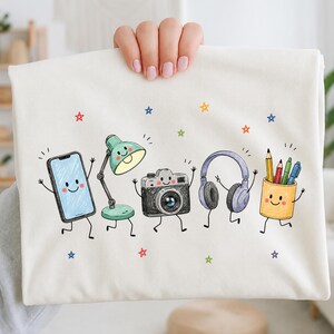 May include: A white t-shirt featuring a colorful graphic print of five anthropomorphic objects: a smartphone, a desk lamp, a camera, headphones, and a pencil holder filled with pens, all surrounded by small, hand-drawn multicolored stars.