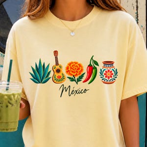 May include: A pale yellow t-shirt featuring a graphic print of an agave plant, a guitar, a marigold flower, a red and green chili pepper, and a decorative vase. Below the illustrations, the word México is written in a black cursive script.