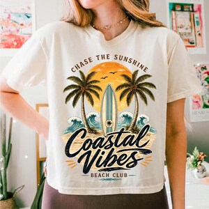 May include: Cream-colored t-shirt with a beach-themed graphic. The design features a surfboard, palm trees, waves, and the text "Chase the Sunshine" and "Coastal Vibes Beach Club". The graphic is in shades of orange, blue, and black.