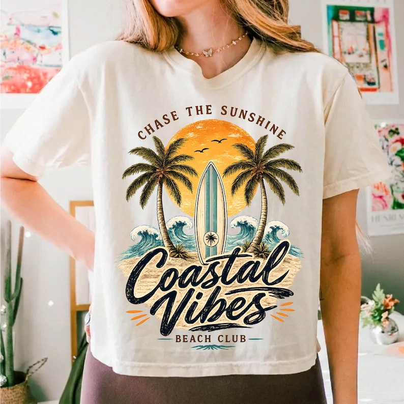 May include: Cream-colored t-shirt with a beach-themed graphic. The design features a surfboard, palm trees, waves, and the text "Chase the Sunshine" and "Coastal Vibes Beach Club". The graphic is in shades of orange, blue, and black.