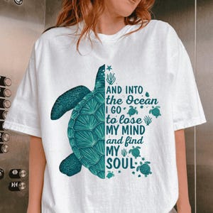 Puede incluir: Camiseta blanca con un diseño de tortuga turquesa en el lado izquierdo y el texto "AND INTO the Ocean I GO to lose MY MIND and find MY SOUL" en el lado derecho. El diseño incluye pequeñas ilustraciones de tortugas.