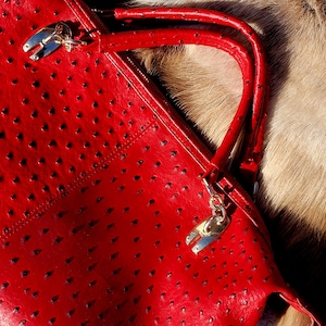 May include: A vibrant red handbag with a textured surface and small perforations. The bag features a top handle and silver-toned hardware. The bag is set against a backdrop of fur.