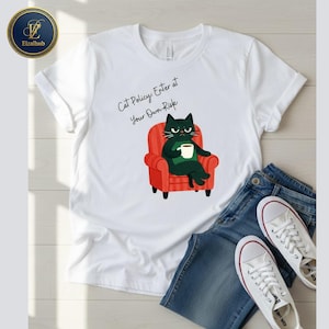 May include: White t-shirt with a graphic of a black cat sitting in a red armchair, holding a coffee cup. The text "Cat Policy: Enter at Your Own Risk" is above the cat. The shirt is paired with blue jeans and white sneakers.