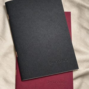 May include: Two staple-bound notebooks stacked on top of each other. The top notebook has a matte black cover, and the bottom notebook has a matte burgundy cover. Both feature the embossed text INKFORGE on the lower right corner.