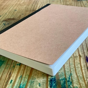 May include: A rectangular notebook with a light brown cover and a black spine. The pages are white, and the edges are also white. The notebook is resting on a wooden surface.