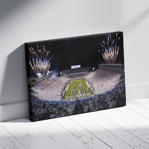 May include: Canvas print of a packed stadium at night, with fireworks bursting in the sky. The field is lit, and the stands are filled with spectators. The canvas is mounted on a wooden frame.