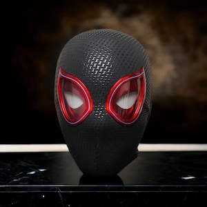 Op de afbeelding: Een zwart Spider-Man-masker met rood omrande ooglenzen. Het masker heeft een gestructureerd, geweven patroon en een strak, nauwsluitend ontwerp. De ooglenzen zijn helderrood met een wit gaasinterieur.