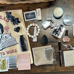 May include: An assortment of items including stickers, a small black purse, a canvas tote bag with text and a graphic, a beaded bracelet, a makeup sponge, and pens, arranged on a wooden surface.