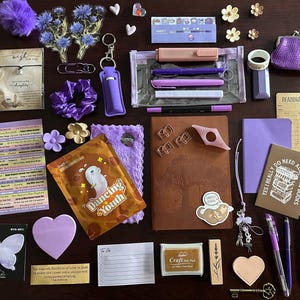 Puede incluir: Una colección de artículos de papelería y accesorios morados, que incluyen bolígrafos, un cuaderno, pegatinas, un monedero y una goma para el pelo. Los artículos están dispuestos sobre una superficie marrón oscuro, creando una presentación visualmente atractiva.