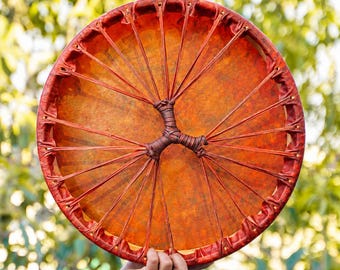 Puede incluir: Un tambor de marco circular con un parche de color naranja rojizo. El parche está sujeto al marco mediante cordones finos y tensos que irradian desde un nudo central envuelto, creando un patrón en forma de estrella en la superficie.