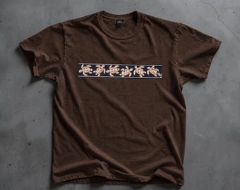 May include: A dark brown short-sleeved t-shirt featuring a horizontal rectangular graphic across the chest. The graphic contains a repeating pattern of light-coloured sea turtles arranged in a row against a dark blue background.