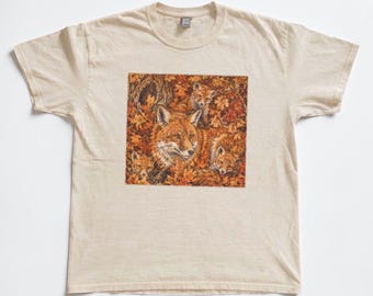 May include: A cream-coloured short-sleeve t-shirt featuring a square graphic print on the chest. The illustration depicts a group of orange foxes nestled among a dense pile of fallen autumn leaves in shades of brown, gold, and deep red.