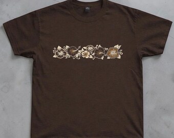 May include: A dark brown short-sleeved t-shirt featuring a horizontal band of hibiscus flowers across the chest. The floral graphic is rendered in shades of cream, light brown, and dark brown, creating a tonal design on the solid brown fabric.