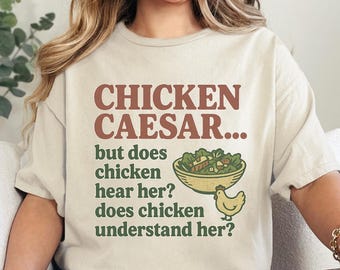May include: A cream-colored short-sleeve t-shirt featuring the brown text CHICKEN CAESAR... and green text but does chicken hear her? does chicken understand her? alongside a graphic illustration of a salad bowl and a small yellow chicken.