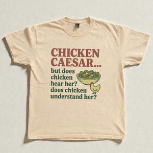 May include: A light beige t-shirt with the text "CHICKEN CAESAR..." in maroon. Below the text is a graphic of a salad bowl and a chicken, with the text "but does chicken hear her? does chicken understand her?"