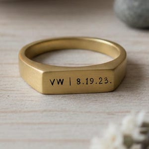 May include: A gold-toned metal signet ring with a flat rectangular face featuring the engraved text VW | 8.19.23. The ring has a smooth, matte finish and a simple band design, presented as a singular piece of jewelry with clear, legible lettering.