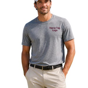 May include: A heather gray short-sleeved t-shirt featuring the text Name Tag Logo in dark red on the left chest, paired with a white baseball cap, a black belt with a gold buckle, and light beige trousers against a plain white background.