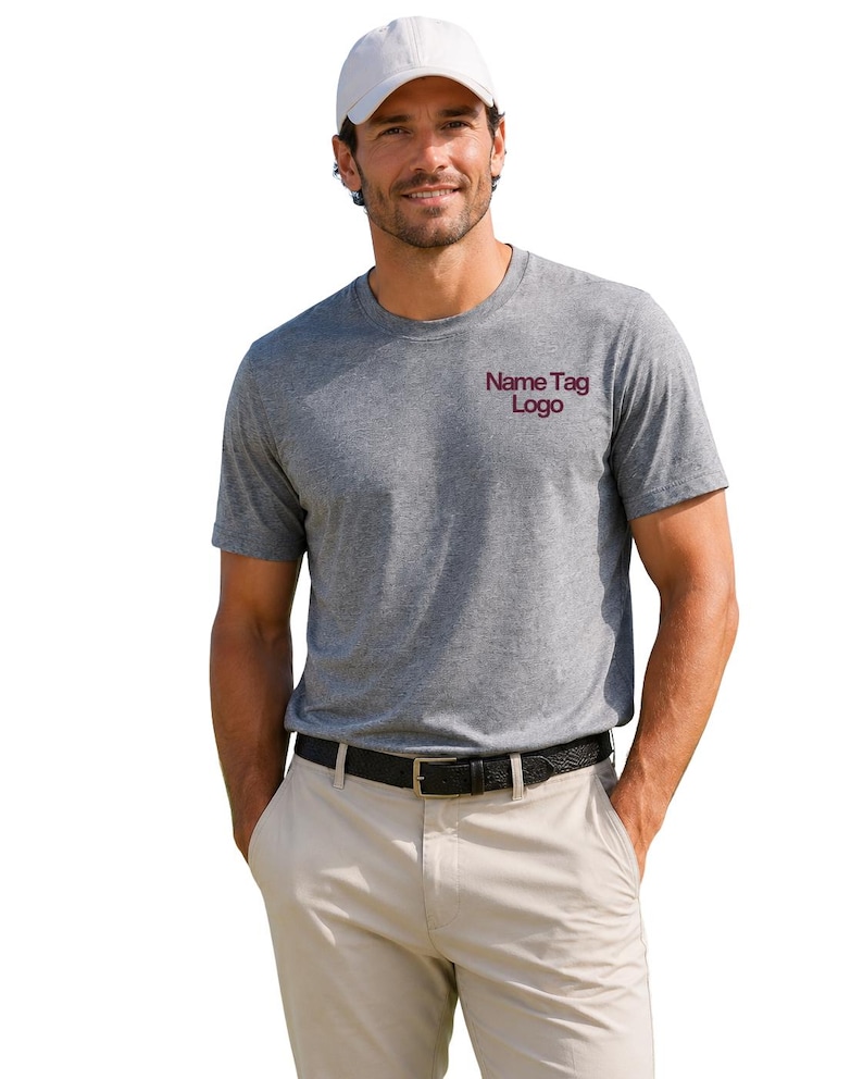 May include: A heather gray short-sleeved t-shirt featuring the text Name Tag Logo in dark red on the left chest, paired with a white baseball cap, a black belt with a gold buckle, and light beige trousers against a plain white background.