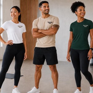 May include: Three short-sleeved crew neck t-shirts in white, beige, and dark green, each featuring the text Your Logo Here on the left chest. The shirts are paired with black athletic leggings and black athletic shorts, creating a coordinated set.