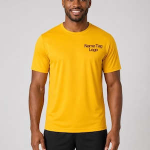May include: A mustard yellow short-sleeved crew neck t-shirt featuring the text "Name Tag Logo" in dark red on the upper left chest, paired with plain black athletic shorts. The shirt has a smooth, lightweight fabric texture and a standard fit.