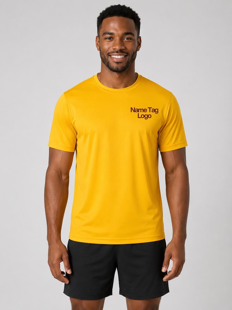 May include: A mustard yellow short-sleeved crew neck t-shirt featuring the text "Name Tag Logo" in dark red on the upper left chest, paired with plain black athletic shorts. The shirt has a smooth, lightweight fabric texture and a standard fit.