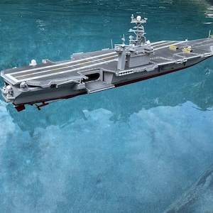 May include: A gray model aircraft carrier floating on water, featuring a flat flight deck with yellow markings, a tall island superstructure, and a dark red hull section visible below the waterline with a small propeller and rudder assembly.