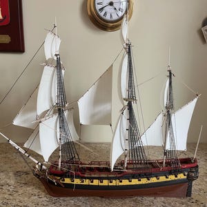 May include: A detailed model ship with white sails, black rigging, and a brown hull with yellow and red accents. The ship has multiple masts and is set against a neutral background. A decorative item.