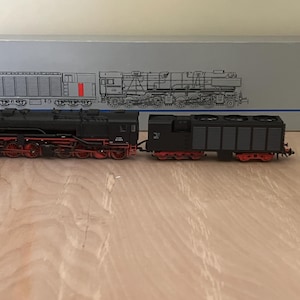 May include: A black model steam locomotive with red wheels and a matching tender, positioned in front of its original gray Märklin digital HO packaging box. The box features a line drawing of the train and the model number 37021 printed on it.