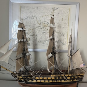 Pode incluir: Modelo detalhado de um navio alto com vários mastros e velas. O navio é marrom e preto com detalhes amarelos, e as velas são bege. Um mapa antigo está no fundo, emoldurado em prata.