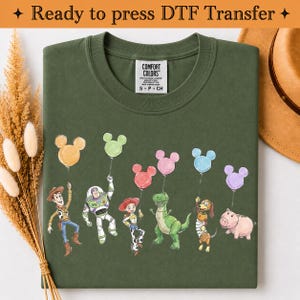 Puede incluir: Una camiseta verde oliva con un estampado gr&aacute;fico de seis personajes de dibujos animados de Toy Story, cada uno sosteniendo un globo con forma de Mickey Mouse de varios colores. La camiseta tiene una etiqueta Comfort Colors en el cuello.