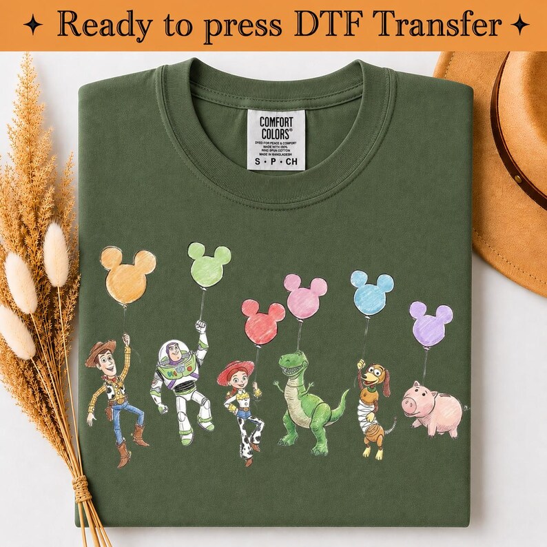 Puede incluir: Una camiseta verde oliva con un estampado gr&aacute;fico de seis personajes de dibujos animados de Toy Story, cada uno sosteniendo un globo con forma de Mickey Mouse de varios colores. La camiseta tiene una etiqueta Comfort Colors en el cuello.