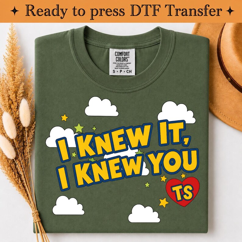 May include: An olive green t-shirt featuring a graphic print with white clouds, yellow stars, and the text "I KNEW IT, I KNEW YOU" in yellow with blue outlines. A red heart contains the yellow letters "TS". The shirt has a Comfort Colors label.