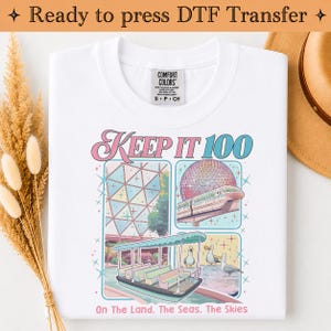 May include: A white t-shirt featuring a graphic print with the text "KEEP IT 100" and "On the Land, The Seas, The Skies". The design includes illustrations of a geodesic dome, a monorail, a boat, and seagulls, accented with small star shapes.