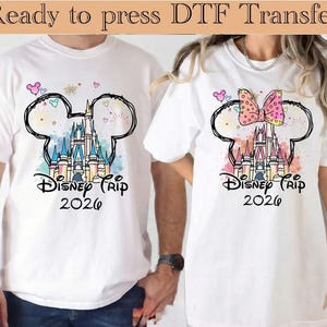 May include: Two white t-shirts featuring a graphic of a castle inside a Mickey Mouse ear outline. The design includes the text Disney Trip 2026, colorful watercolor splashes, and fireworks. The right shirt features a pink polka dot bow.