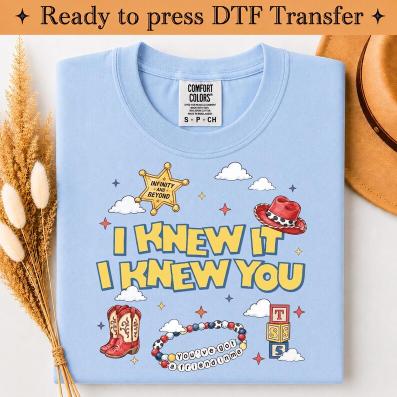 May include: A light blue t-shirt featuring a graphic print with the text I KNEW IT I KNEW YOU, a yellow sheriff star with INFINITY AND BEYOND, a red cowboy hat, a pair of red cowboy boots, a beaded bracelet, and a stack of three lettered blocks.