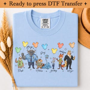 Puede incluir: Una camiseta azul claro con un estampado gr&aacute;fico de varios personajes animados, cada uno sosteniendo un globo de color con forma de cabeza de rat&oacute;n. Los personajes est&aacute;n etiquetados con nombres como Flash, Bogo, Gary, Nibbles, Judy Hopps, Nick Wilde y Mayor.