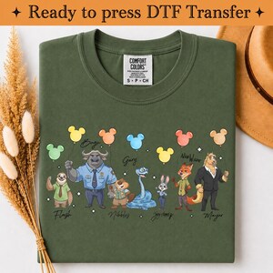 Puede incluir: Una camiseta verde oliva con un estampado gr&aacute;fico de siete personajes animados de Zootopia, cada uno sosteniendo un globo de color. Los personajes est&aacute;n etiquetados con sus nombres: Flash, Bogo, Nibbles, Gary, Judy Hopps, Nick Wilde y Mayor.