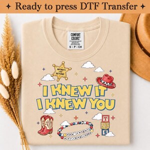 May include: A beige t-shirt featuring a graphic print with the text I KNEW IT I KNEW YOU. The design includes a yellow sheriff star with INFINITY AND BEYOND, a red cowboy hat, red and white boots, a beaded bracelet with YOU'VE GOT A FRIEND IN ME, and blocks.