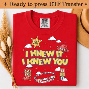 May include: A red t-shirt featuring a graphic print with the text I KNEW IT I KNEW YOU. The design includes a yellow sheriff star, a red cowboy hat, a pair of red cowboy boots, a colorful beaded bracelet, and a stack of three lettered blocks.
