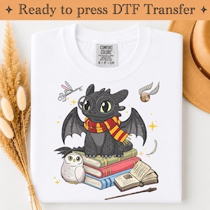 Op de afbeelding: Een wit t-shirt met een afbeelding van een zwarte draak met een ronde bril en een rood-geel gestreepte sjaal, zittend op een stapel boeken naast een witte uil, een vliegende sleutel, een gouden snaai, een open boek en een toverstaf.