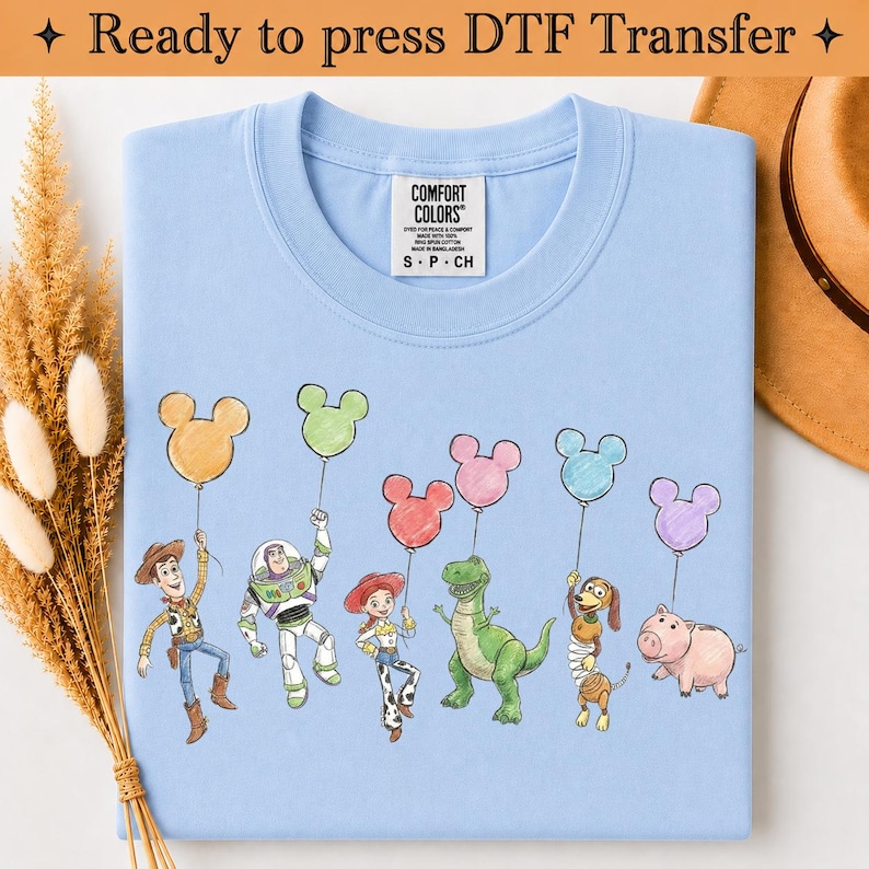 Puede incluir: Una camiseta azul claro con un estampado gr&aacute;fico de seis personajes animados de Toy Story, cada uno sosteniendo un globo con forma de Mickey Mouse de varios colores. La camiseta tiene una etiqueta de la marca Comfort Colors en el cuello y el texto Ready to press DTF Transfer.