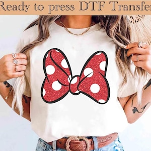 Könnte beinhalten: Weißes T-Shirt mit einer großen, roten Glitzer-Schleife mit weißen Punkten und schwarzem Umriss. Der Text "Ready to press DTF Transfer" steht oben. Das T-Shirt wird von einer Person getragen.