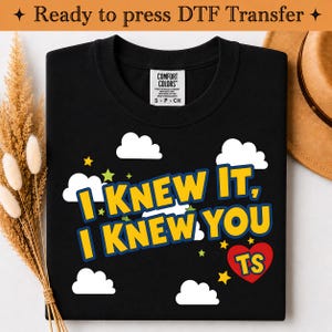 May include: A black t-shirt featuring a graphic print with white clouds, yellow stars, and the text "I KNEW IT, I KNEW YOU" in yellow with a blue outline. A red heart contains the yellow letters "TS". The shirt has a Comfort Colors brand label.